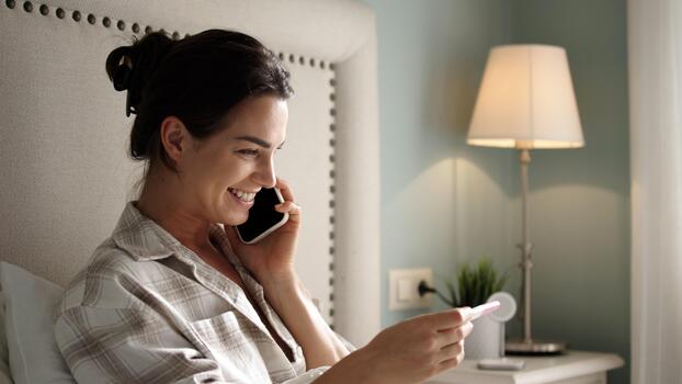 woman-in-cozy-bedroom-smiling-while-holding-pregnancy-test-and-talking-on-phone-showcasing-joyful-moment-of-anticipation-and-excitement-for-future-family-photo